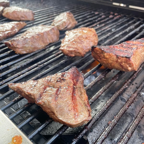 Several cuts of Bailey's Smokehouse BBQ meat grilling on a black grate over flames.