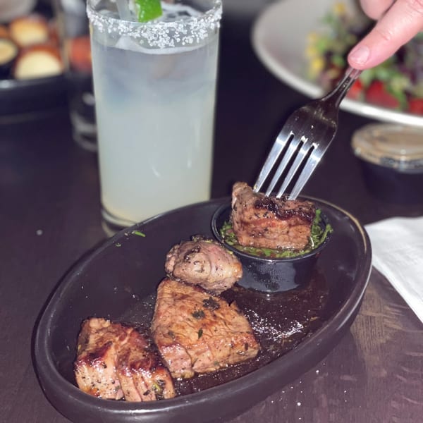 A fork lifts a piece of grilled steak from a black plate next to a salted margarita.