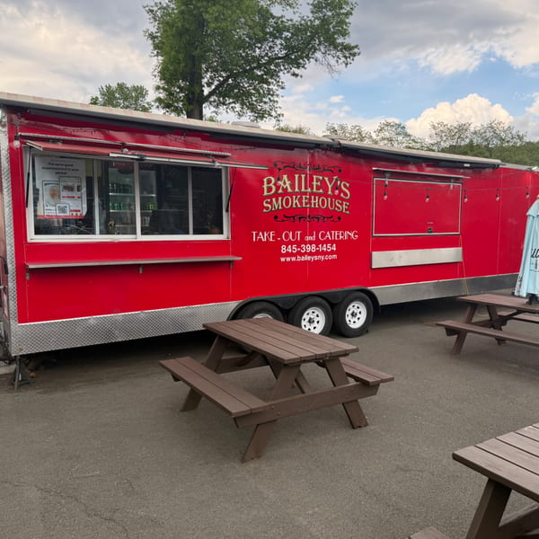 Bailey's Smokehouse red food truck with picnic tables outside under a cloudy sky.