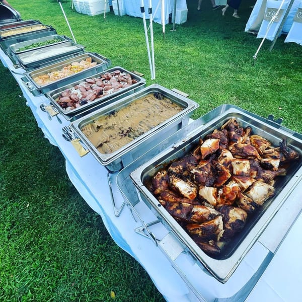 A buffet line of Bailey's Smokehouse BBQ dishes, including chicken, sausage, and sides, on a grassy lawn.