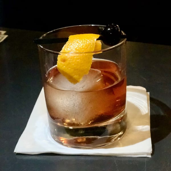 An Old Fashioned cocktail with a large ice cube, lemon peel, and cherry on a white napkin.