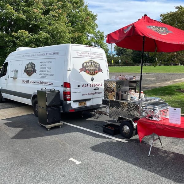 Bailey's Smokehouse catering van and food cart with a red umbrella set up outdoors.