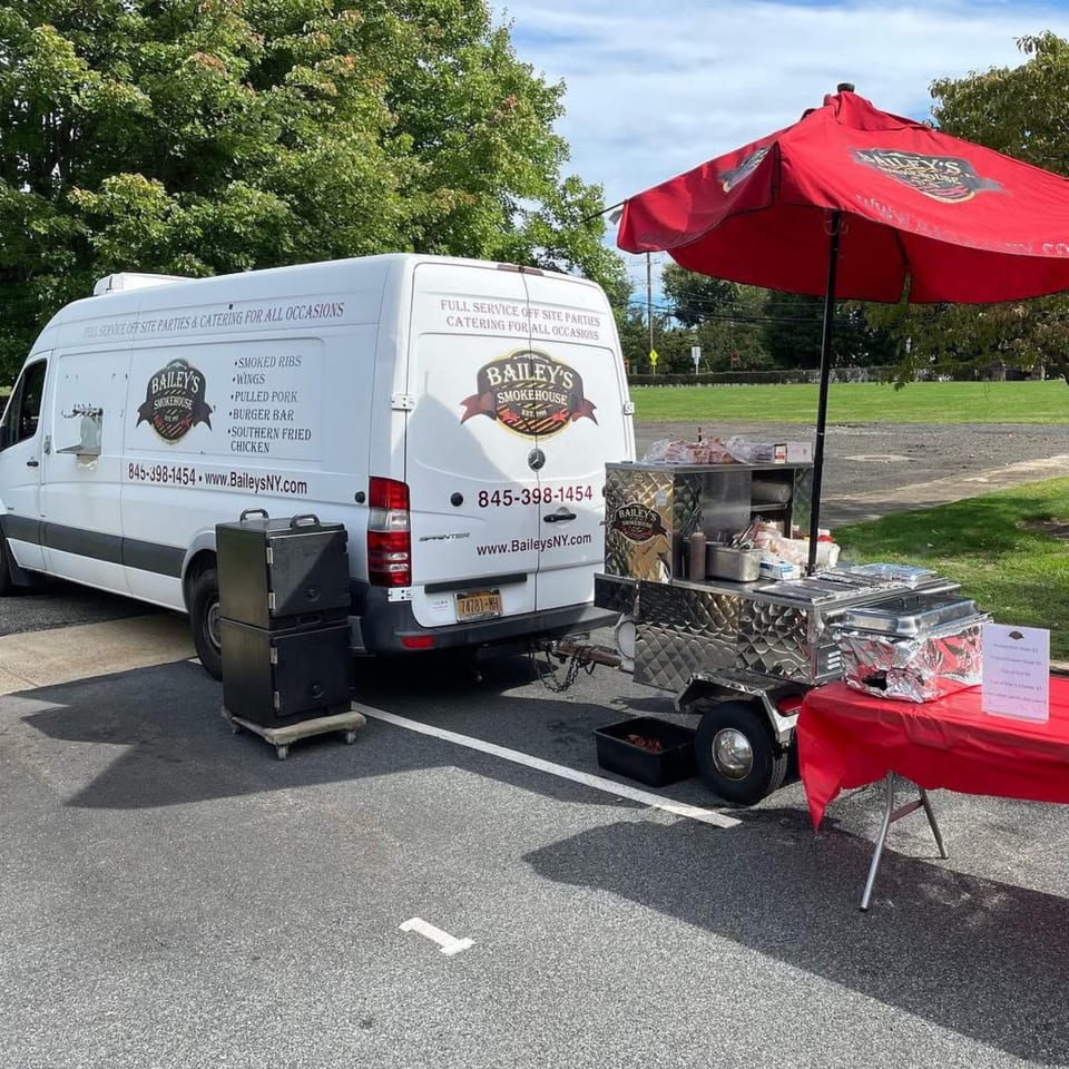 Bailey's Smokehouse catering van and food cart with a red umbrella set up outdoors.