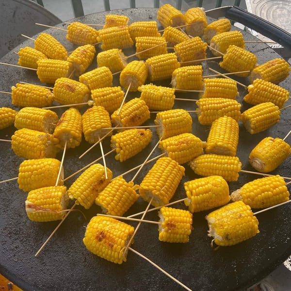 Numerous grilled corn on the cob skewers cooking on a large, round, dark griddle.