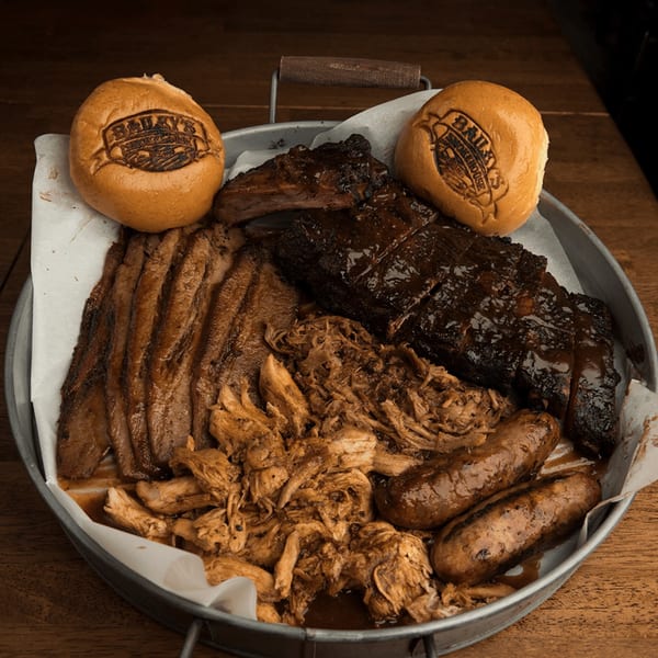 A metal tray overflowing with Bailey's Smokehouse BBQ meats, including brisket, ribs, pulled pork, and sausages, with branded buns.