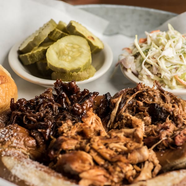 Close-up of a BBQ pulled pork sandwich with pickles and coleslaw on a white paper-lined tray.