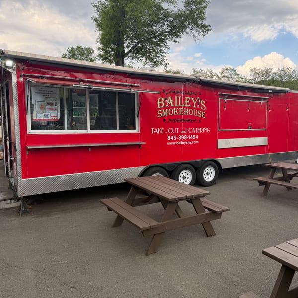 Bailey's Smokehouse red food truck with picnic tables outside under a cloudy sky.