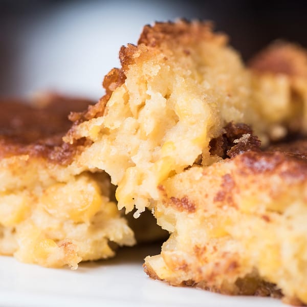 Close-up of golden-brown cornbread with visible corn kernels, broken into pieces on a white plate.