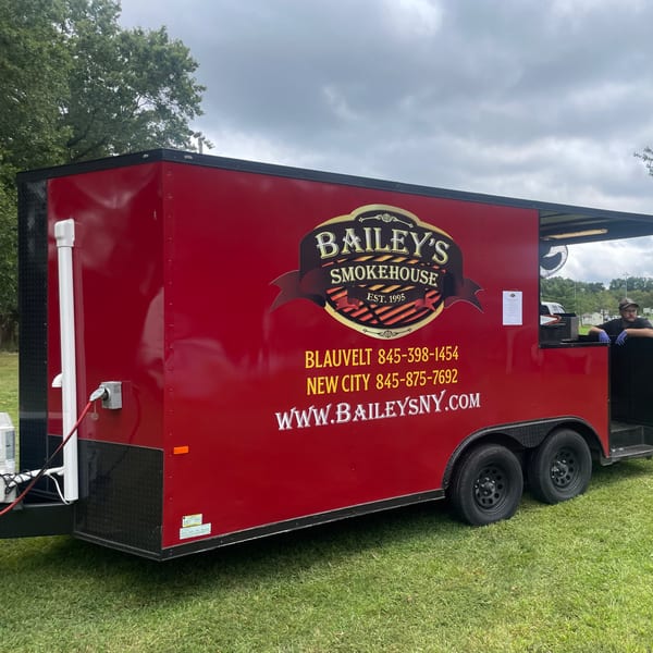 Bailey's Smokehouse red food truck with a person inside, parked on green grass under a cloudy sky.