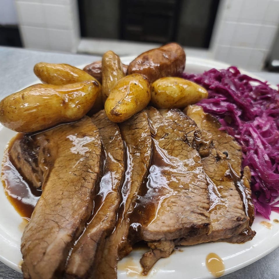 Sliced brisket with gravy, roasted potatoes, and red cabbage on a white plate at Bailey's Smokehouse.