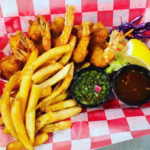 Fried shrimp and french fries with two dipping sauces and a lemon wedge in a red and white checkered basket.