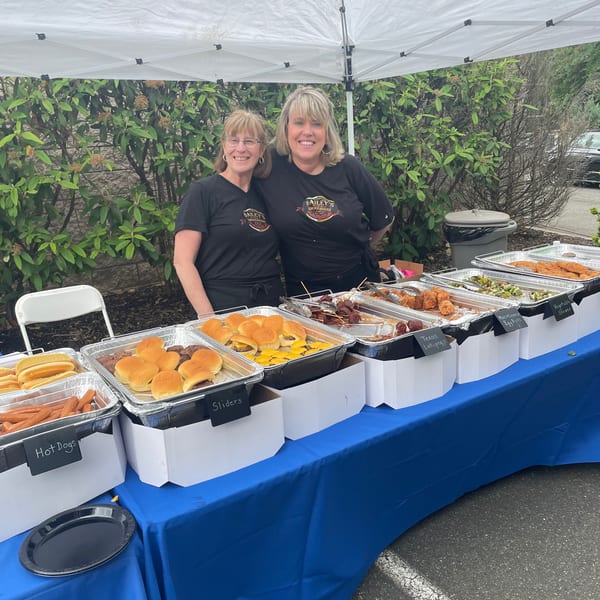 Two women from Bailey's Smokehouse stand behind a buffet of hot dogs, sliders, and other BBQ items.
