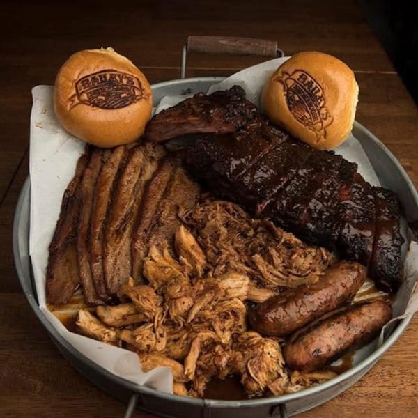 A metal tray filled with Bailey's Smokehouse BBQ brisket, pulled pork, ribs, sausages, and branded buns.
