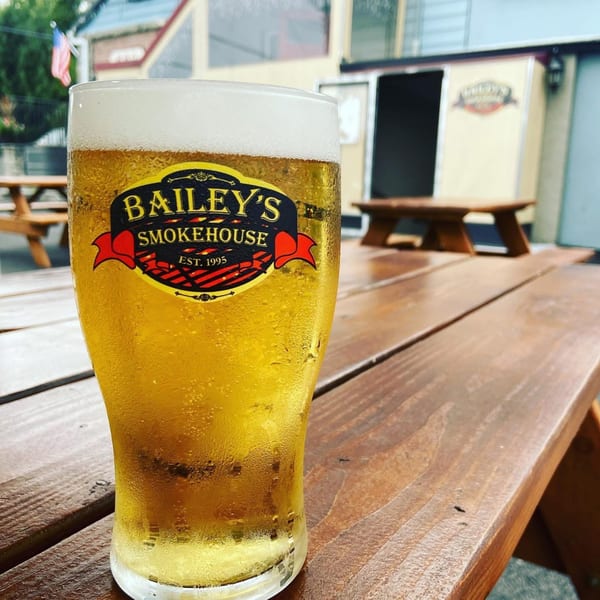 A frosty glass of beer with the Bailey's Smokehouse logo sits on a wooden picnic table.