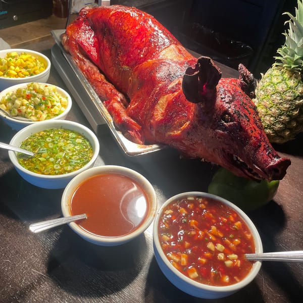 A whole roasted pig, glistening red, surrounded by bowls of sauces and salsas, with a pineapple.