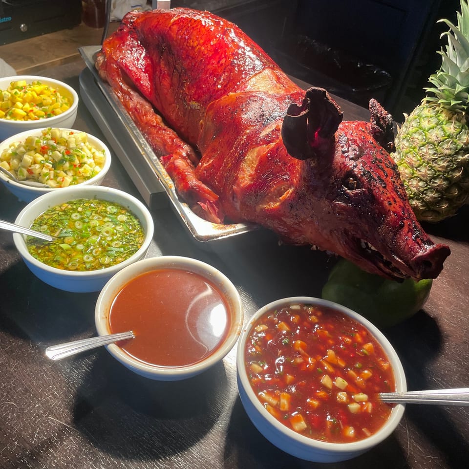 A whole roasted pig, glistening red, surrounded by bowls of sauces and salsas, with a pineapple.