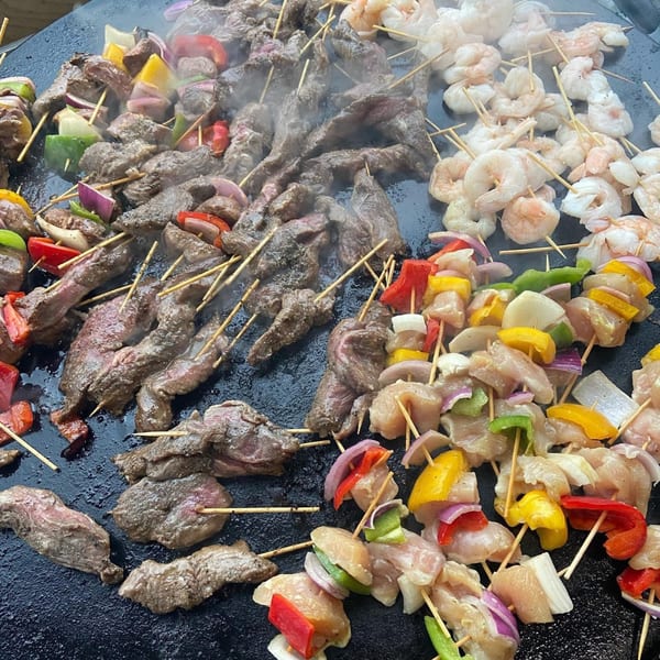 Assorted skewers of beef, chicken, shrimp, and vegetables grilling on a large flat-top surface.