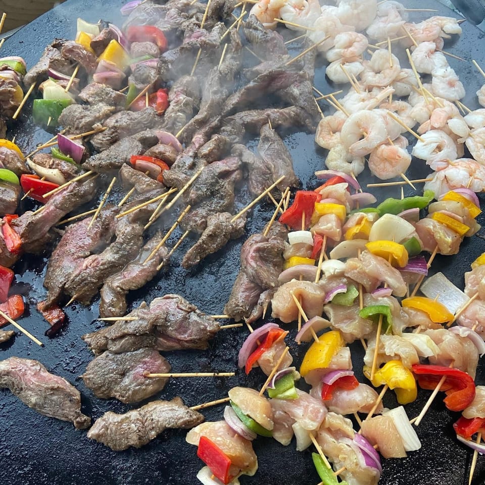 Assorted skewers of beef, chicken, shrimp, and vegetables grilling on a large flat-top surface.
