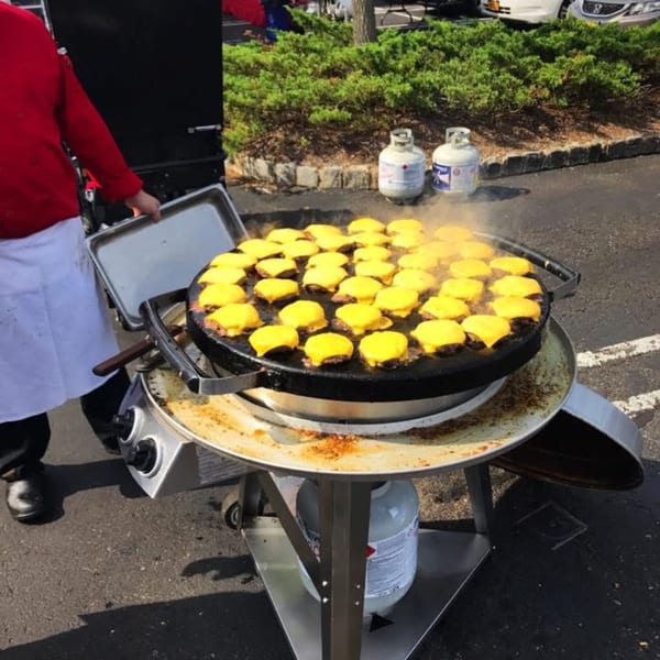 A chef cooks many cheeseburger sliders on a large outdoor griddle, with propane tanks nearby.