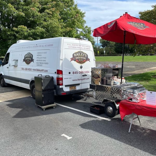 Bailey's Smokehouse catering van and food cart with a red umbrella set up outdoors.