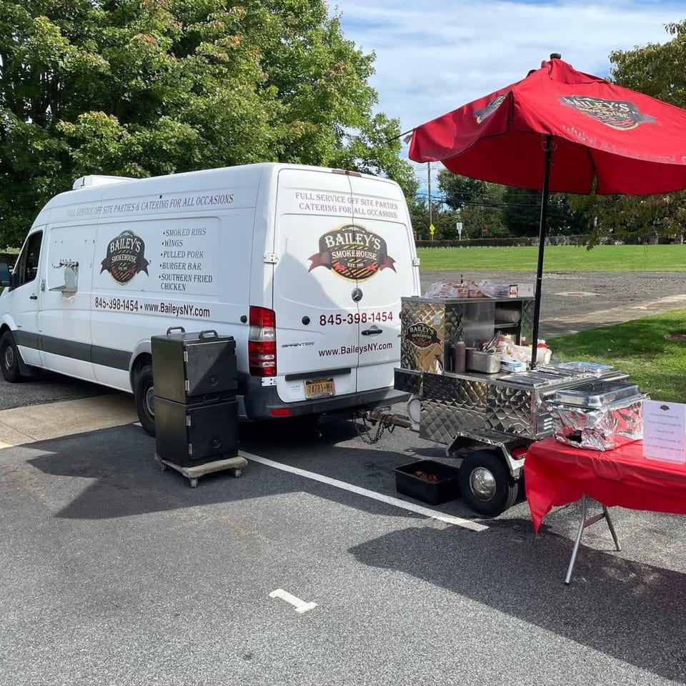 Bailey's Smokehouse catering van and food cart with a red umbrella set up outdoors.